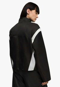 Giacca nera con colletto alto, caratterizzata da accenti diagonali bianchi, texture liscia, vestibilità comoda e dettagli notevoli sulla cucitura posteriore.