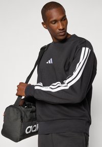Sweatshirt preto com design de três riscas brancas nas mangas e pequeno logotipo. Mala de desporto preta com o nome da marca em branco, segurada na mão direita.