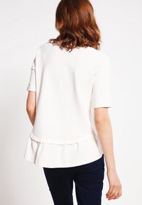 Vit kortärmad topp med texturerad yta, med en utställd peplumslängd, som bärs med mörka byxor. Baksidan är synlig.