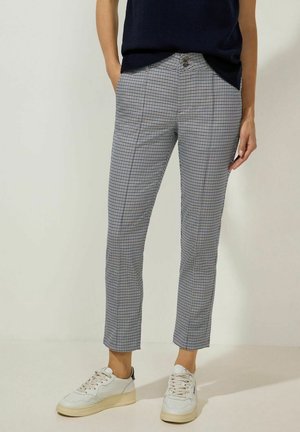 Person iført navyblå top, slanke ternede bukser med forreste læg og hvide sneakers, stående foran en ensartet lys baggrund.