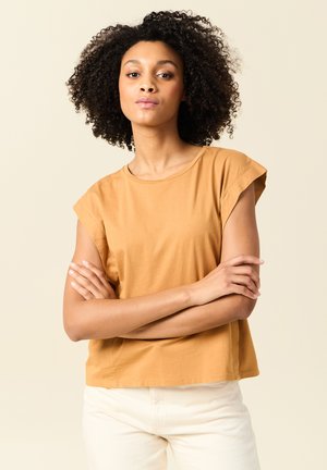 Senf-farbenes, kurzärmeliges T-Shirt aus weicher Baumwolle mit entspannter Passform, rundem Ausschnitt und dezenten Stickdetails, kombiniert mit hellen Hosen.