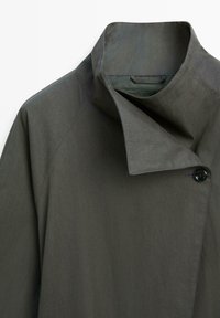 Donkergrijze jas met hoge kraag en asymmetrische voorflap die met een enkele zwarte knoop is afgesloten, met gedetailleerde stiksels op de schouder en kraag.