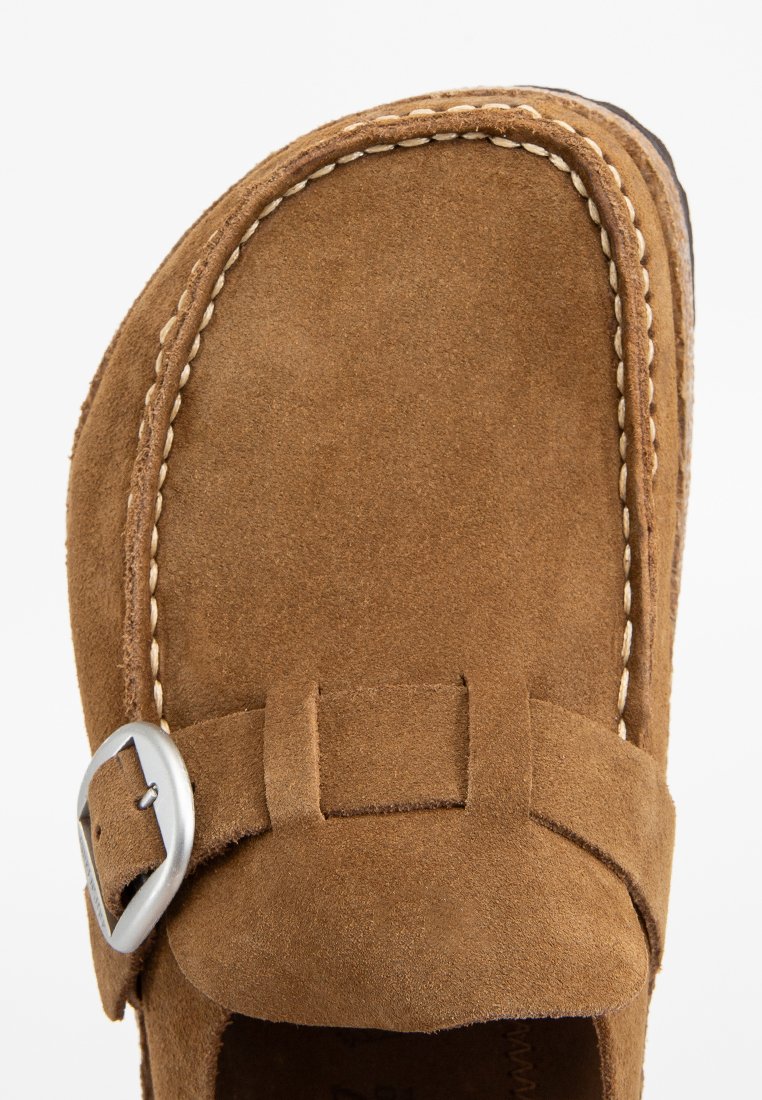 Brązowy zamszowy but wsuwany z okrągłym noskiem, białe przeszycia wzdłuż krawędzi i akcent w postaci srebrnej metalowej klamry. Płaska podeszwa.
