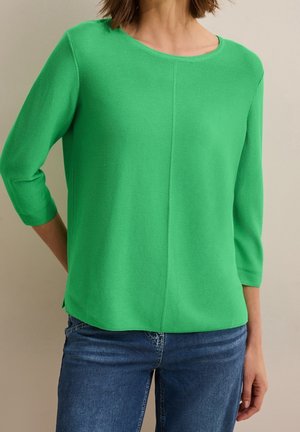 Person wearing a bright green long-sleeve top with a round neckline and blue jeans, standing against a plain beige background.