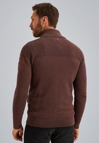 Brązowy sweter z dzianiny z wysokim kołnierzem, z teksturowanej tkaniny, z prążkowanymi mankietami i gładką łatą na lewym ramieniu.