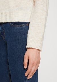 Rechte Hand ruht auf dunkelblauen Denim-Jeans, trägt einen cremefarbenen Strickpullover mit gerippten Bündchen vor einem schlichten Hintergrund.