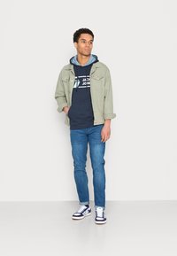 Giacca verde chiaro sopra un hoodie blu navy con logo, jeans blu e sneakers bianche con accenti blu navy. Outfit semplice e casual.