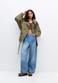 Olijfgroene oversized trenchcoat met zwarte knopen, gelaagd over een bruine crop top en lichtblauwe wijdpijpen denim jeans. Bruine klompen met studs.