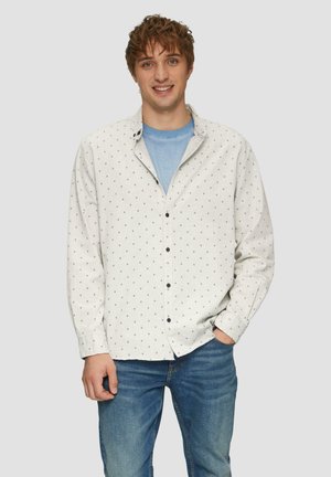 Witte overhemd met kleine zwarte diamantenpatronen, gecombineerd met een lichtblauwe t-shirt eronder. Gekozen met blauwe jeans.