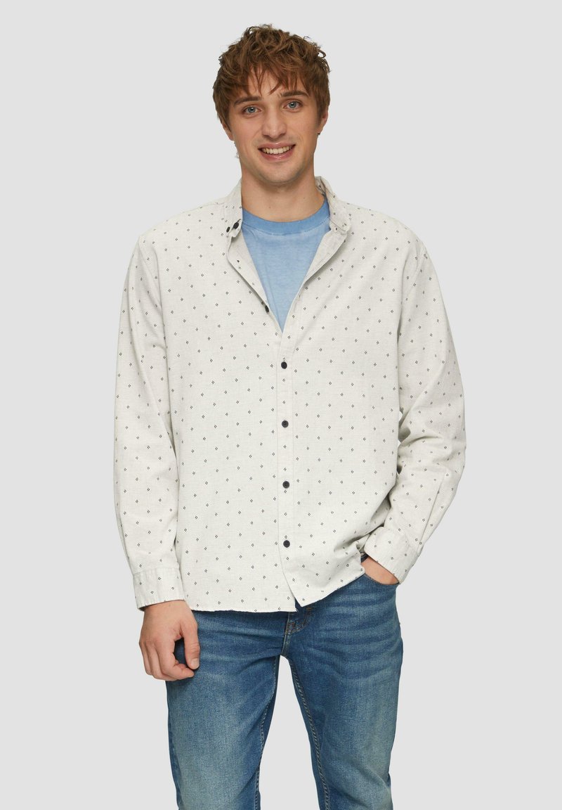 Witte overhemd met kleine zwarte diamantenpatronen, gecombineerd met een lichtblauwe t-shirt eronder. Gekozen met blauwe jeans.