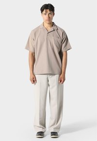 Beżowa koszula z krótkim rękawem i kołnierzykiem, z kieszenią, w połączeniu z jasnymi, luźnymi spodniami i czarnymi butami. Widoczny teksturowany wzór materiału.