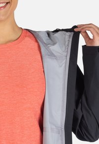 Zwarte jas met een grijze binnenvoering, voorzien van een rits en een hand die de kraag vasthoudt. Heldere koraalkleurige shirt zichtbaar eronder.