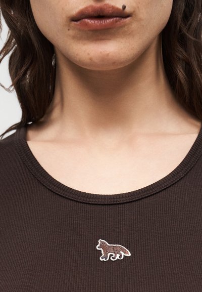 Ορατό το κάτω μέρος του προσώπου και ο λαιμός μιας γυναίκας, που φοράει ένα σκούρο καφέ πλεκτό τοπ με ένα μικρό κεντητό λογότυπο αλεπούς κοντά στον γιακά.