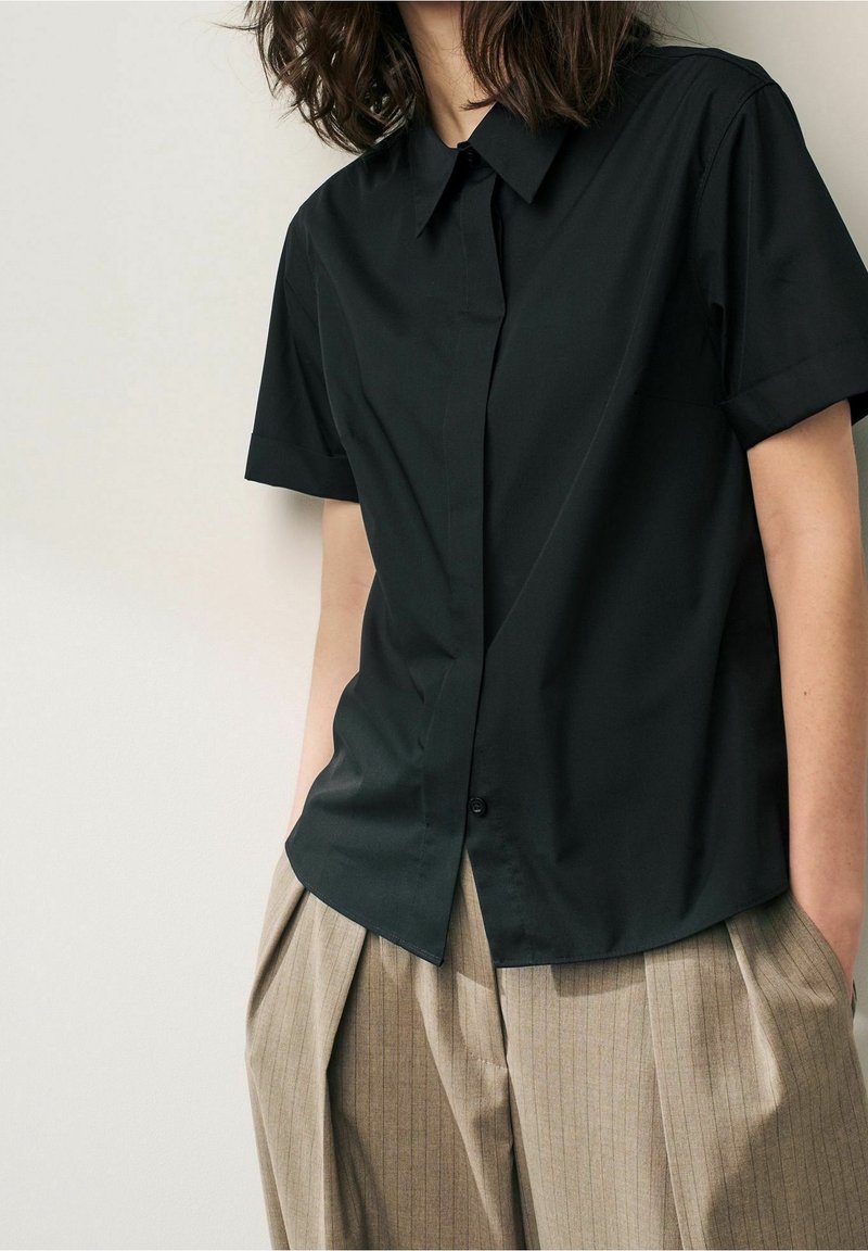 Chemise noire à manches courtes avec boutons et col pointu, présentant une texture lisse et un léger éclat, associée à un pantalon beige à fines rayures.