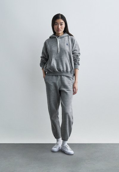 Γυναίκα που φοράει γκρι φούτερ με κουκούλα και αντίστοιχο παντελόνι φόρμας με λευκά αθλητικά παπούτσια, στέκεται μπροστά σε έναν απλό ανοιχτόχρωμο τοίχο.