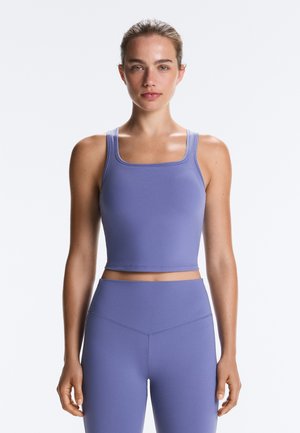 Giovane donna con i capelli raccolti indossa un crop top lilla aderente e leggings a vita alta su uno sfondo bianco neutro.