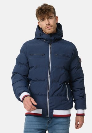 Navy blue puffer jacket with a hood, featuring zippered chest pockets, elasticated cuffs, and contrasting red and white ribbed hem.