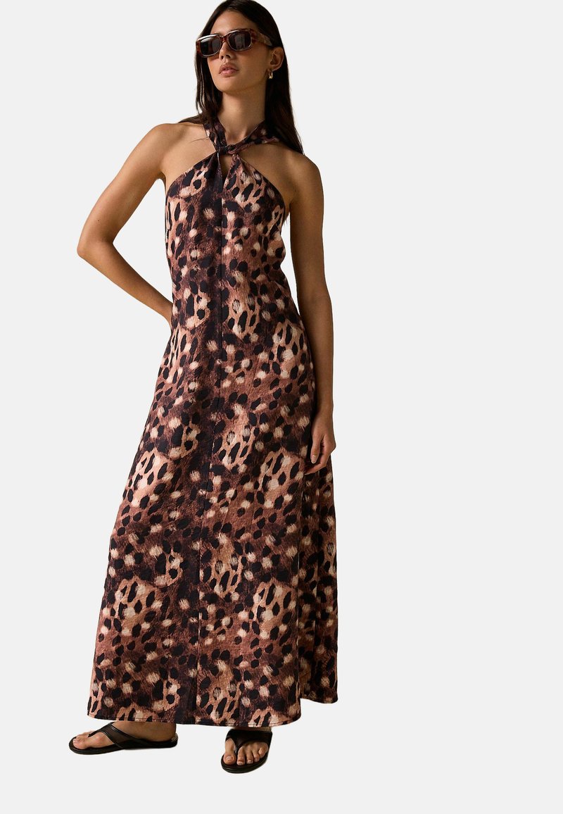 Femme portant une robe longue dos nu à imprimé léopard marron et noir, lunettes de soleil et sandales noires, debout avec une main sur la hanche.