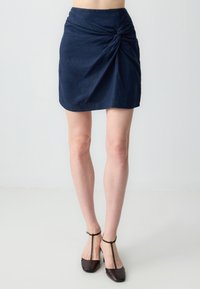 Navyblaue Minirock mit einem verdrehten Knotendesign an der Taille, aus einem strukturierten Stoff gefertigt. Kombiniert mit dunkelbraunen Ankle-Strap-Pumps.