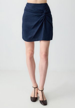 Mini-jupe bleu marine avec un design de nœud torsadé à la taille, fabriquée dans un tissu texturé. Assortie à des talons à brides de cheville brun foncé.