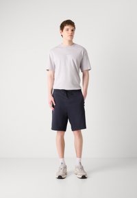 Grå t-shirt med kort ärm och logotyp, tillsammans med svarta knälånga shorts med fickor. Modellen bär ljusa sneakers med vita strumpor.