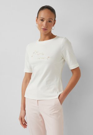 Vrouw met naar achteren gekamd haar, draagt grote oorbellen, een wit shirt met korte mouwen en cursieve tekst, en lichtroze broek, staande tegen een grijze achtergrond.