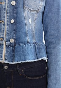 Ljusblå denimjacka med fransad nederkant, framfickor, silverfärgade knappar och en peplumruffle vid midjan. Bar över mörka jeans.