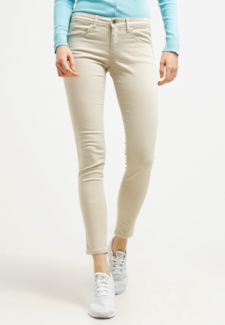 Jeans ajustados beige con bolsillos frontales y cierre de botón. Combinados con zapatillas deportivas grises. La tela parece suave con un ligero estiramiento.