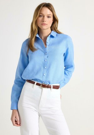 Kvinde iført en lyseblå langærmet skjorte med knapper, stoppet ind i højtaljede hvide bukser med et brunt bælte.