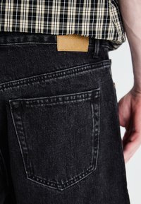 Sorte denim jeans med en baglomme, der har et brunt lædermærke. En ternet skjorte dækker delvist taljen. Overordnet mat tekstur.