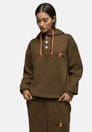 Femme portant un sweat à capuche marron et un pantalon assorti avec des cordons orange et des poches à boutons, regardant vers le bas avec les mains près de la taille.