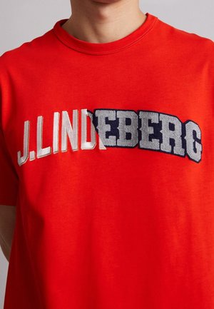 Person trägt ein hellrotes T-Shirt mit weiß-grau gemischtem besticktem Schriftzug „J.LINDEBERG“ auf der Brust.