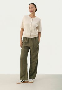Cardigan court beige clair à manches courtes avec boutons, associé à un pantalon large olive, et des baskets grises avec des bandes noires.