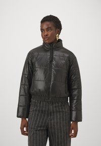 Doudoune noire avec col montant, fermeture éclair, sections matelassées et taille élastique. Le tissu a une texture brillante et lisse.