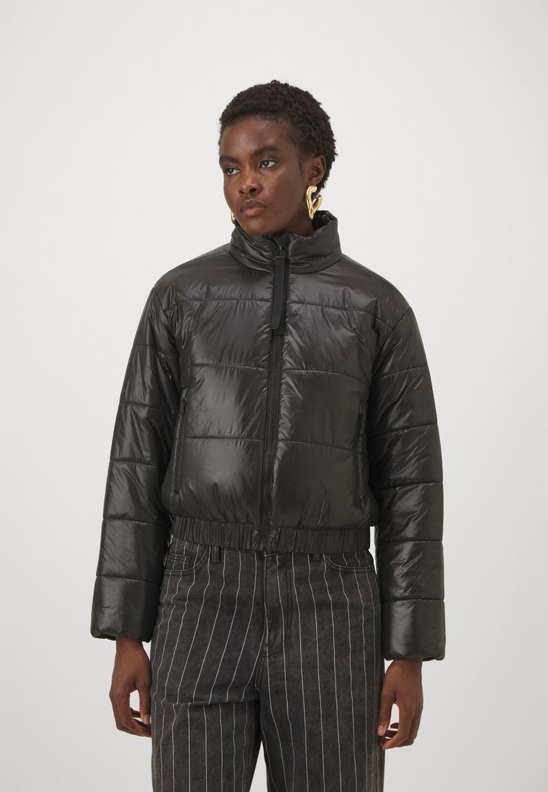 Doudoune noire avec col montant, fermeture éclair, sections matelassées et taille élastique. Le tissu a une texture brillante et lisse.