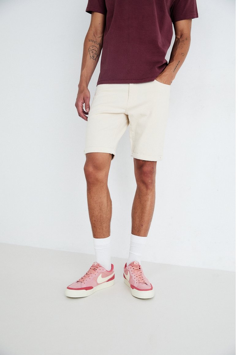 Homme portant un short blanc, un t-shirt bordeaux, des chaussettes blanches et des baskets roses et blanches, debout avec une main dans la poche.