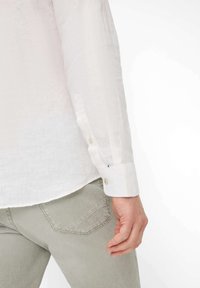 Witte linnen overhemd met een knoopmanchet, gecombineerd met lichtgrijze broek. Opvallende details zijn de knoopsluiting en een subtiele textuur.