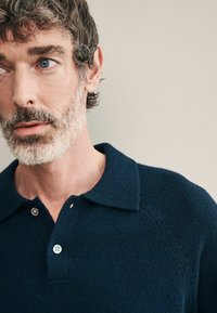 Sötétkék kötött póló, texturált anyagból, bordázott gallérral, két gombos választóval és raglan ujjal.