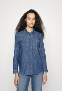 Camisa de denim azul com mangas longas, fecho de botão, colarinho e um bolso no peito. O tecido tem um acabamento texturizado e um corte reto.