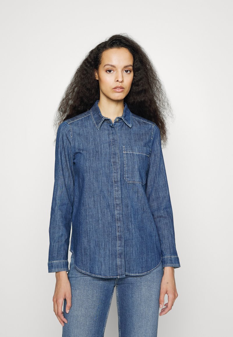 Camisa de denim azul com mangas longas, fecho de botão, colarinho e um bolso no peito. O tecido tem um acabamento texturizado e um corte reto.