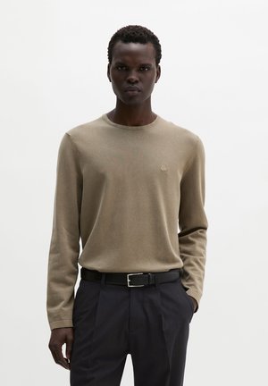 Mann trägt ein beiges Langarmhemd, in schwarze Hose mit schwarzem Gürtel gesteckt, und steht vor einem schlichten weißen Hintergrund.