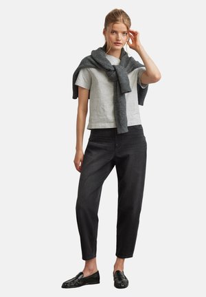 Junge Frau, die ein hellgraues T-Shirt, einen dunkelgrauen Pullover über den Schultern, schwarze Hosen und schwarze Loafers trägt, steht vor einem einfarbigen Hintergrund.