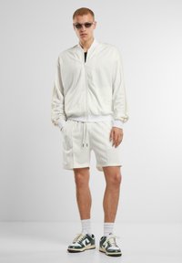 Giacca bomber bianca e pantaloni corti abbinati, entrambi in tessuto liscio. Sneakers con dettagli verdi. Modella con occhiali da sole e calze corte.
