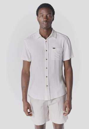 Joven de pie, mirando hacia adelante, vestido con una camisa blanca de manga corta abotonada y pantalones cortos beige, frente a un fondo claro y liso.