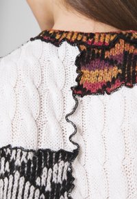 Neulepaita, jossa on kermaista väriä olevia puikko- ja palmikkokuvioita, ja jossa on värikäs monivärinen röyhelökaulus oranssina, mustana ja purppurana, sekä aaltoilevat reunat.