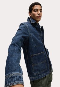 Chaqueta de mezclilla en azul oscuro, con cuello alto, bolsillos frontales y cierre de botones. Tejido texturizado con detalle de costura visible.