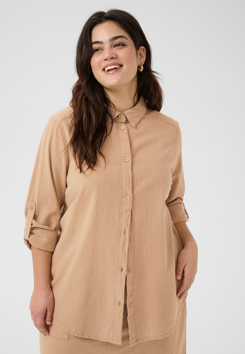 Camisa de botones marrón claro con una textura suave, mangas enrolladas, cuello y bolsillos frontales. Diseño casual para un uso versátil.