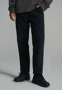 Czarne, lekkie syntetyczne spodnie o luźnym kroju, z elastycznym pasem i delikatną fakturą. W zestawieniu z czarnymi sznurowanymi butami.