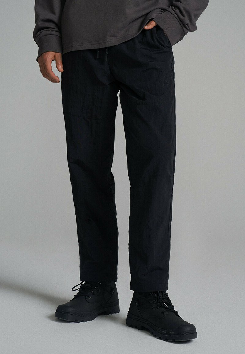 Czarne, lekkie syntetyczne spodnie o luźnym kroju, z elastycznym pasem i delikatną fakturą. W zestawieniu z czarnymi sznurowanymi butami.