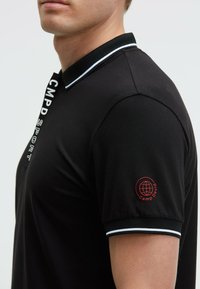 Camisa polo negra con cuello y acentos de manga a rayas blancas. Presenta texto vertical "CMPD SPORT" y un logo rojo en la manga izquierda.